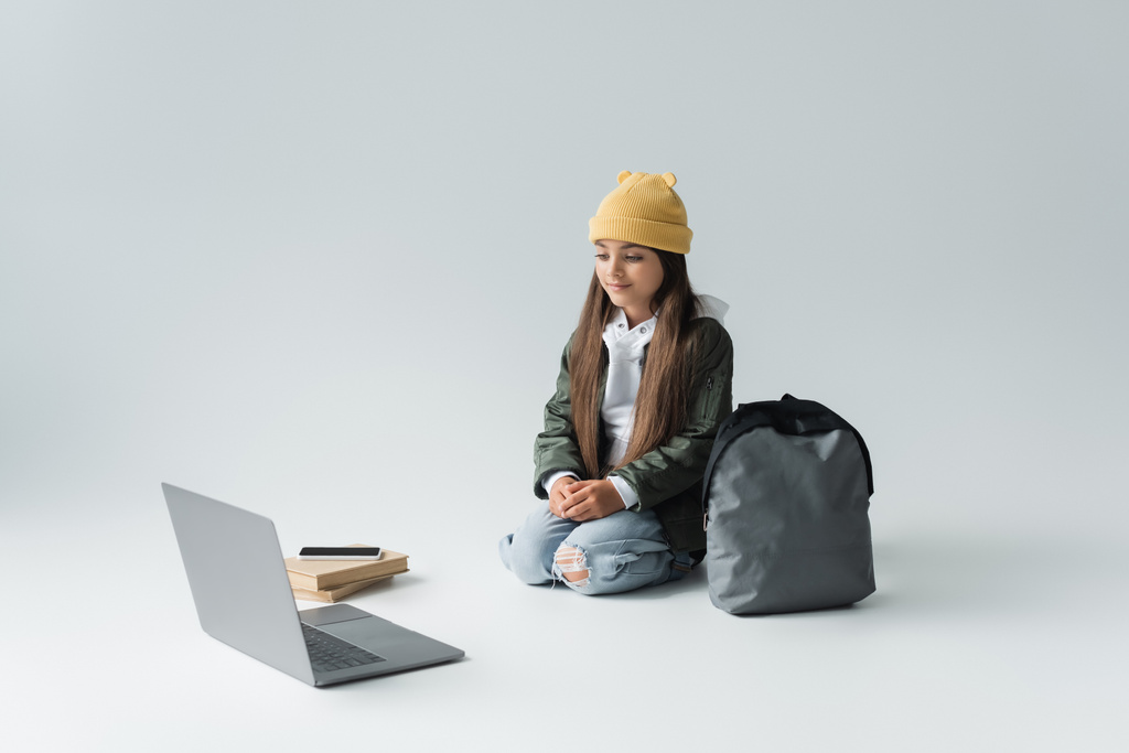 full length of adorable girl in trendy autumnal outfit looking at laptop while sitting near smartphone on books and backpack on grey - Foto, imagen