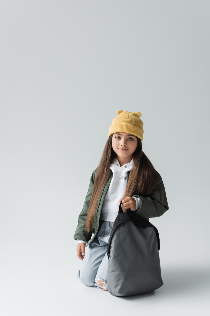 full length of cheerful kid in trendy autumnal outfit and beanie hat holding backpack on grey - Photo, image