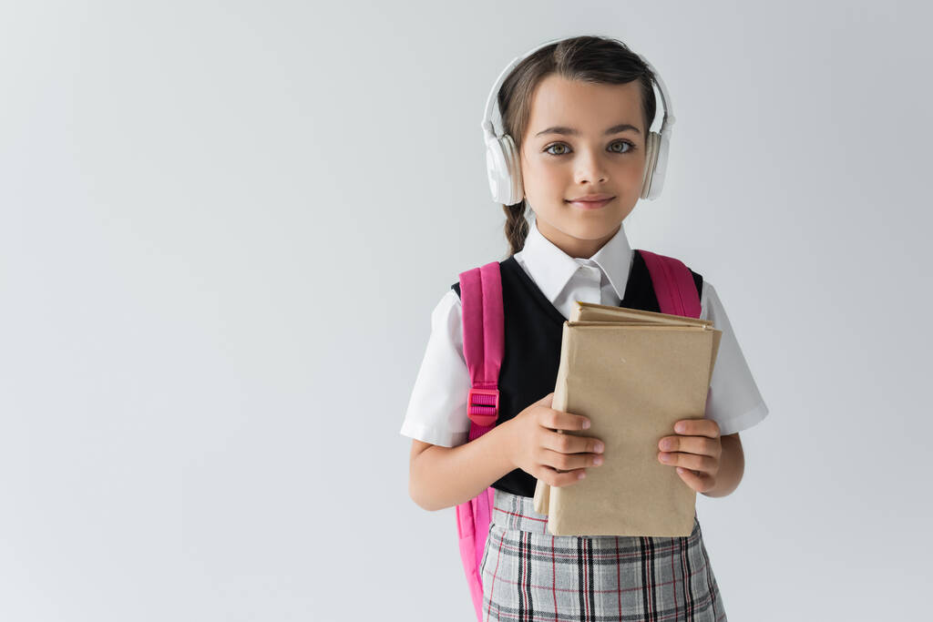 girl in school uniform and wireless headphones holding books isolated on grey - Foto, Bild