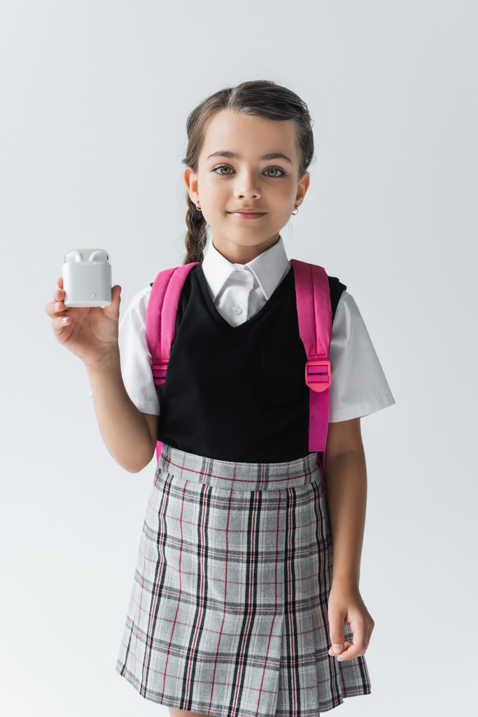 весела школярка в уніформі стоїть з рюкзаком і тримає футляр для навушників ізольовано на сірому
 - Фото, зображення