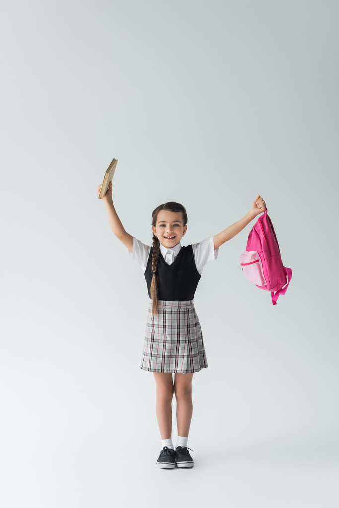 full length of excited schoolgirl in uniform holding pink backpack and book on grey  - Valokuva, kuva