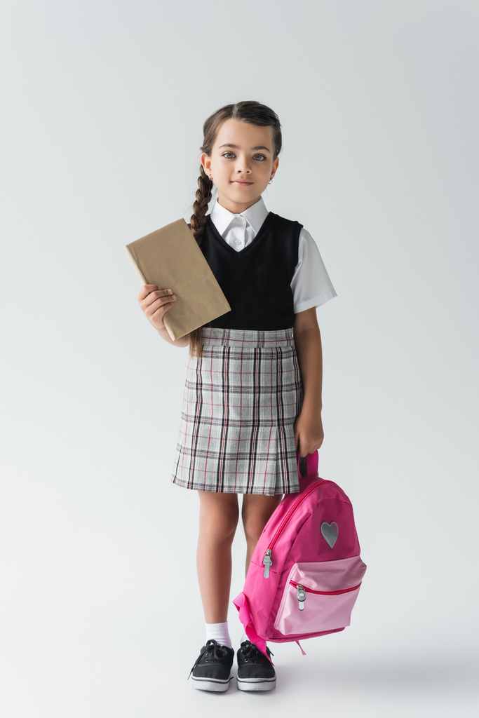 full length of adorable schoolgirl in uniform holding pink backpack and book on grey  - Valokuva, kuva