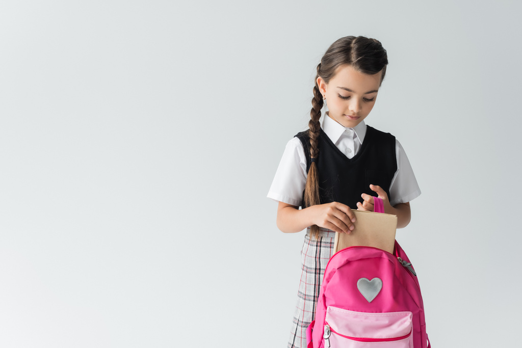 cute schoolgirl in uniform putting book in pink backpack isolated on grey - 写真・画像