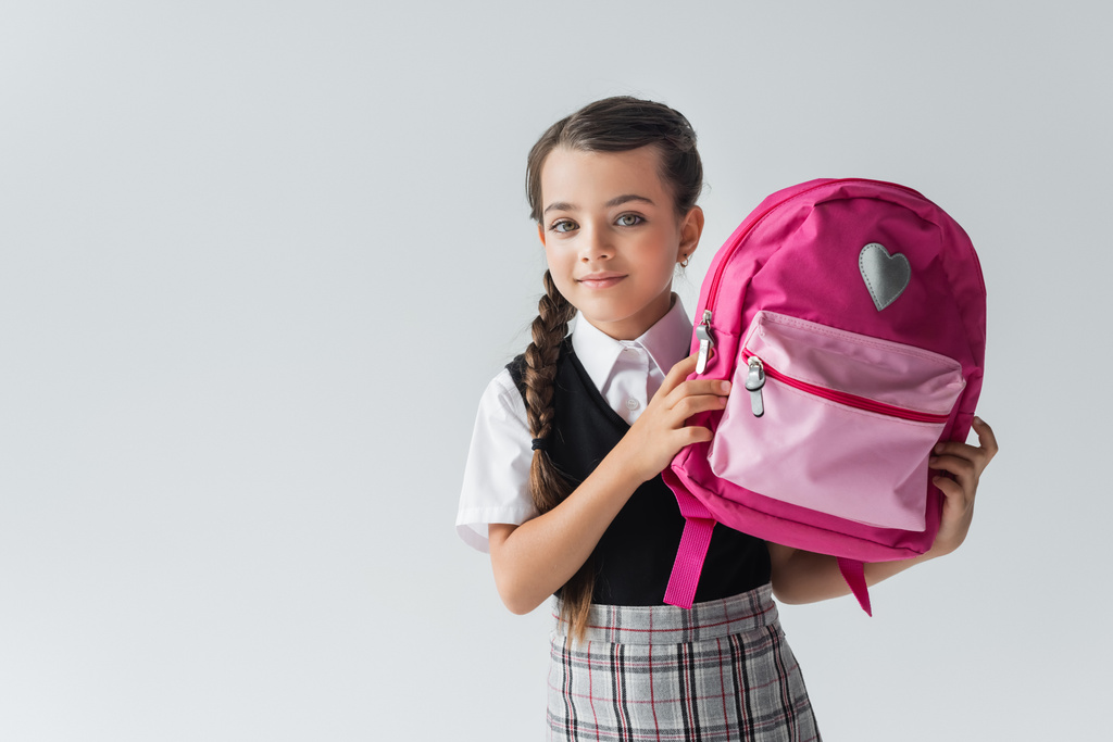 cute schoolgirl in uniform holding pink backpack and smiling isolated on grey - Fotografie, Obrázek
