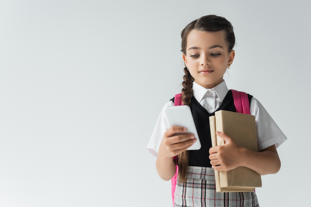 adorable schoolgirl in uniform using smartphone and holding books isolated on grey - Photo, image
