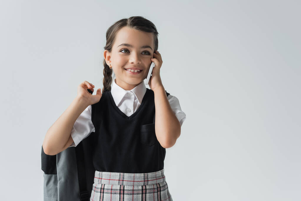 happy schoolgirl holding backpack and talking on smartphone isolated on grey - Foto, Imagem