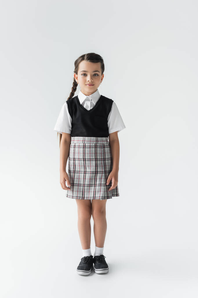 full length of cute schoolgirl in uniform standing and looking at camera on grey - Valokuva, kuva