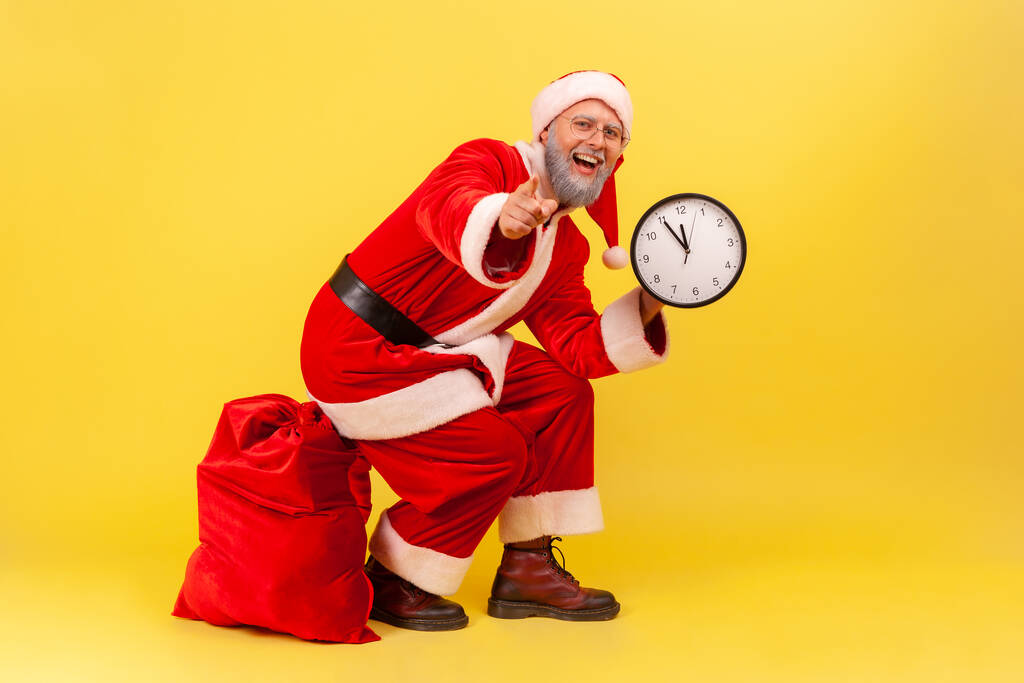 Щасливий позитивний літній чоловік з сірою бородою в костюмі Санта-Клауса сидить на червоній сумці з подарунками, тримаючи настінний годинник в руках і вказуючи на камеру. Внутрішній студійний знімок ізольовано на жовтому фоні
 - Фото, зображення