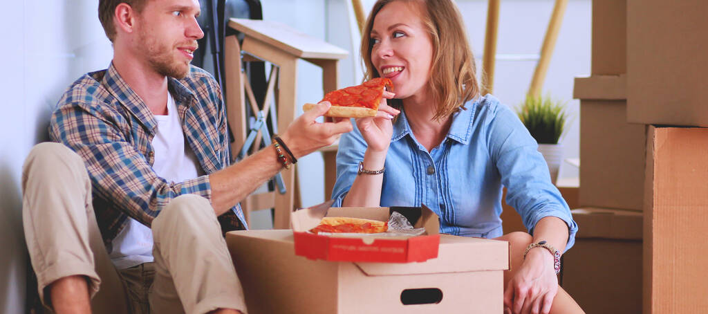 Ένα νεαρό ζευγάρι κάνει ένα διάλειμμα για πίτσα στο πάτωμα αφού μετακομίσει σε ένα νέο σπίτι με κουτιά γύρω του. Νεαρό ζευγάρι. - Φωτογραφία, εικόνα