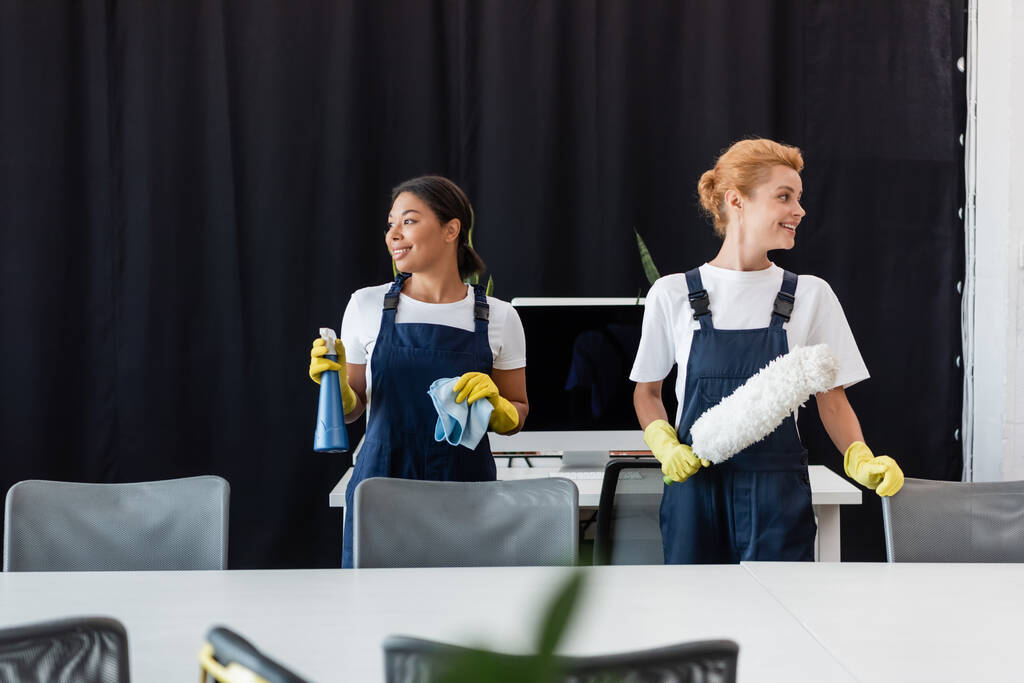 межрасовые женщины в комбинезоне, держащие чистящие средства и улыбающиеся в офисе - Фото, изображение