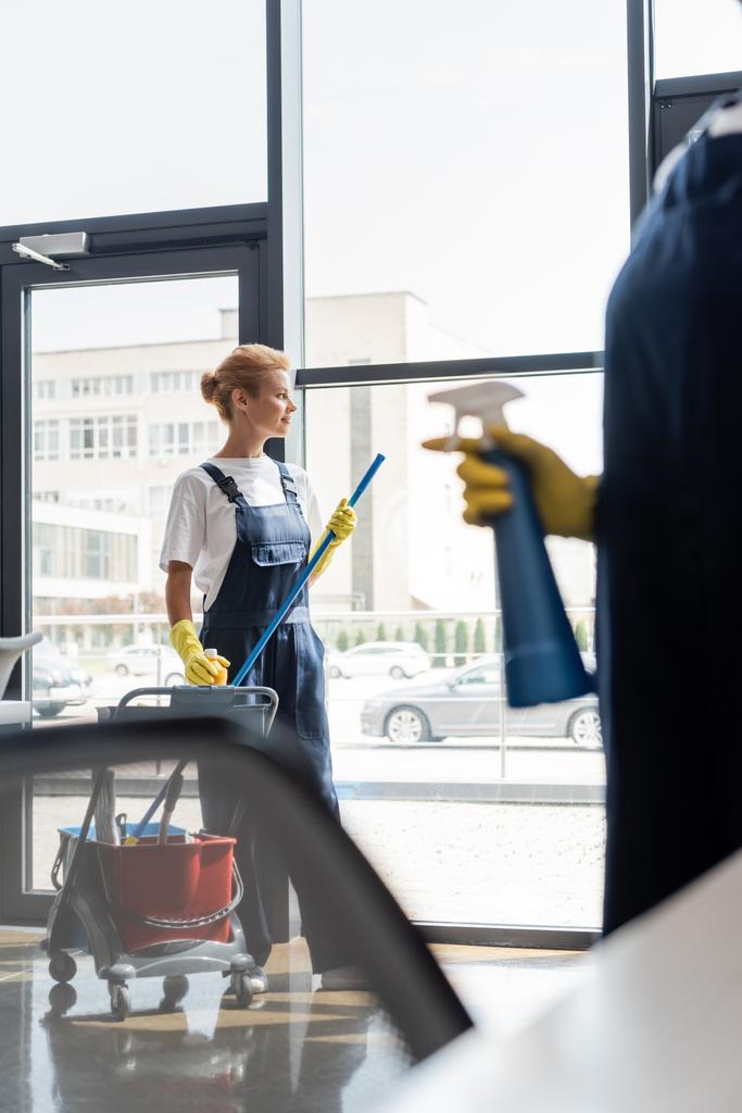 женщина возле тележки с чистящими средствами и коллега с моющим средством на размытом переднем плане - Фото, изображение