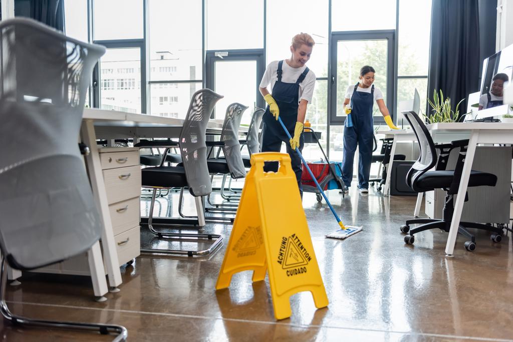woman in overalls washing floor with mop near attention board and bi-racial colleague - Photo, Image