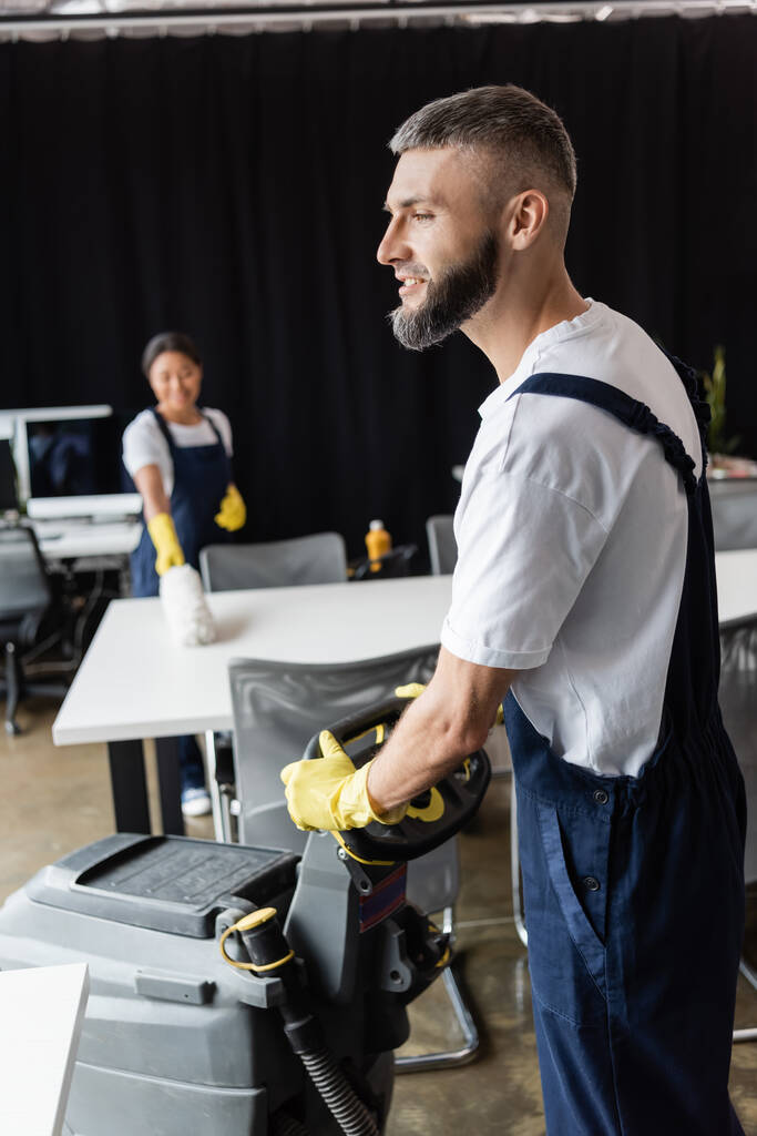 hombre barbudo con máquina de fregar piso cerca de la mujer bi-racial que limpia el escritorio sobre fondo borroso - Foto, imagen