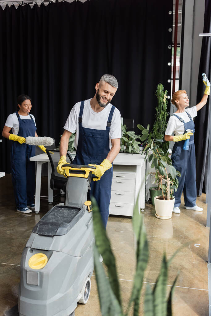 szczęśliwy człowiek obsługujący maszynę do płukania podłogi podczas multietnicznych kobiet sprzątanie biura - Zdjęcie, obraz