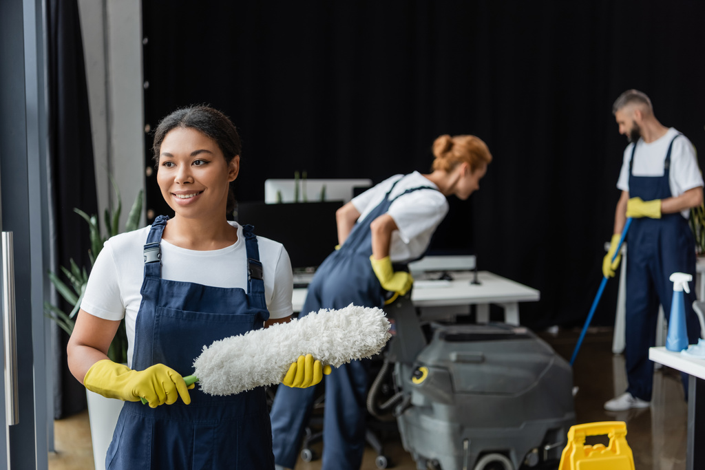 улыбающаяся двурасовая женщина с пыльной щеткой рядом с коллегами и стиральной машиной для пола на размытом фоне - Фото, изображение