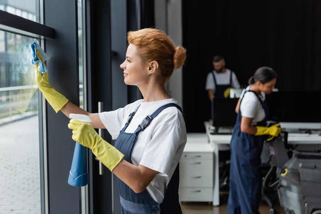 mujer en guantes de goma ventana de limpieza con trapo y detergente cerca borrosa colegas interracial - Foto, Imagen