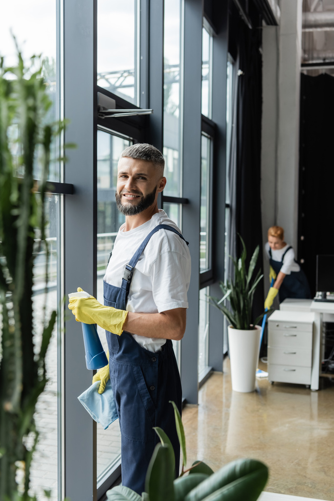 cheerful bearded man with detergent and rag looking at camera near office windows - Photo, Image