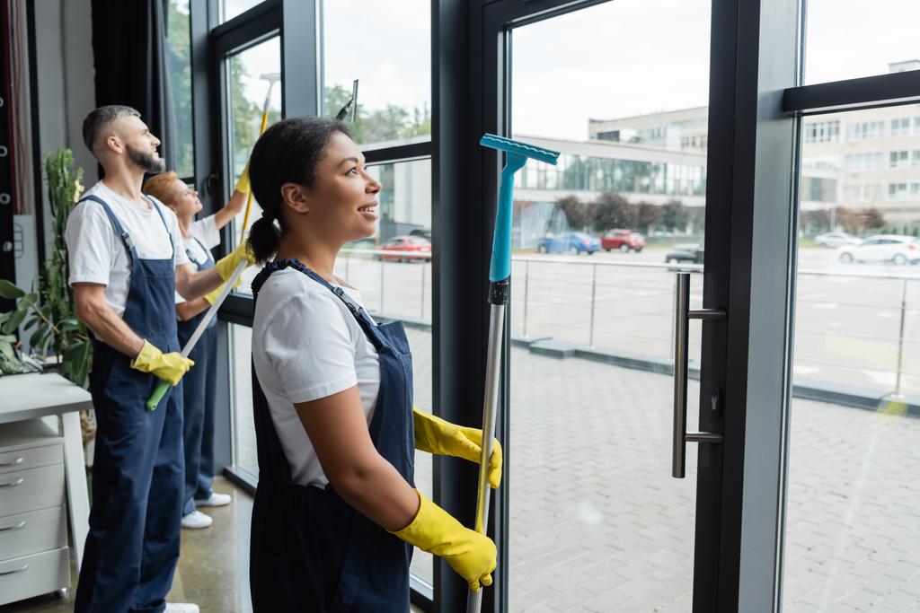 joven bi-racial mujer en ropa de trabajo sonriendo cerca de compañeros de trabajo lavando ventanas de oficina - Foto, imagen