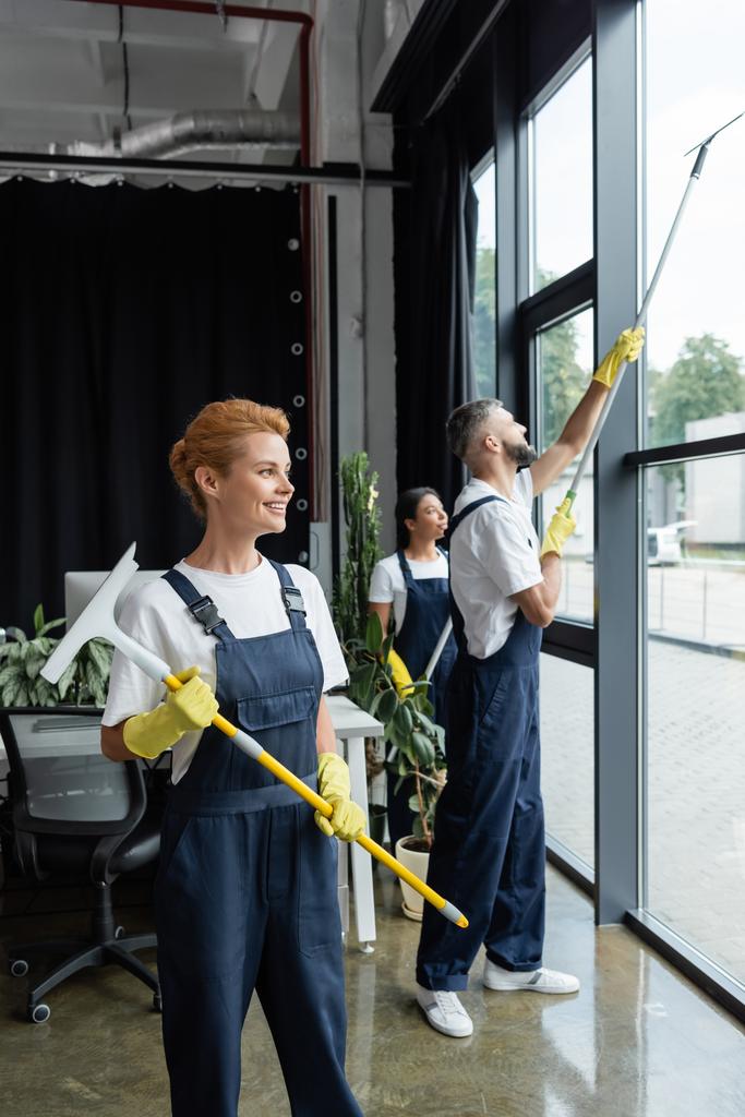glimlachende vrouw in uniform houden venster wisser terwijl interraciale collega 's wassen kantoor ramen - Foto, afbeelding