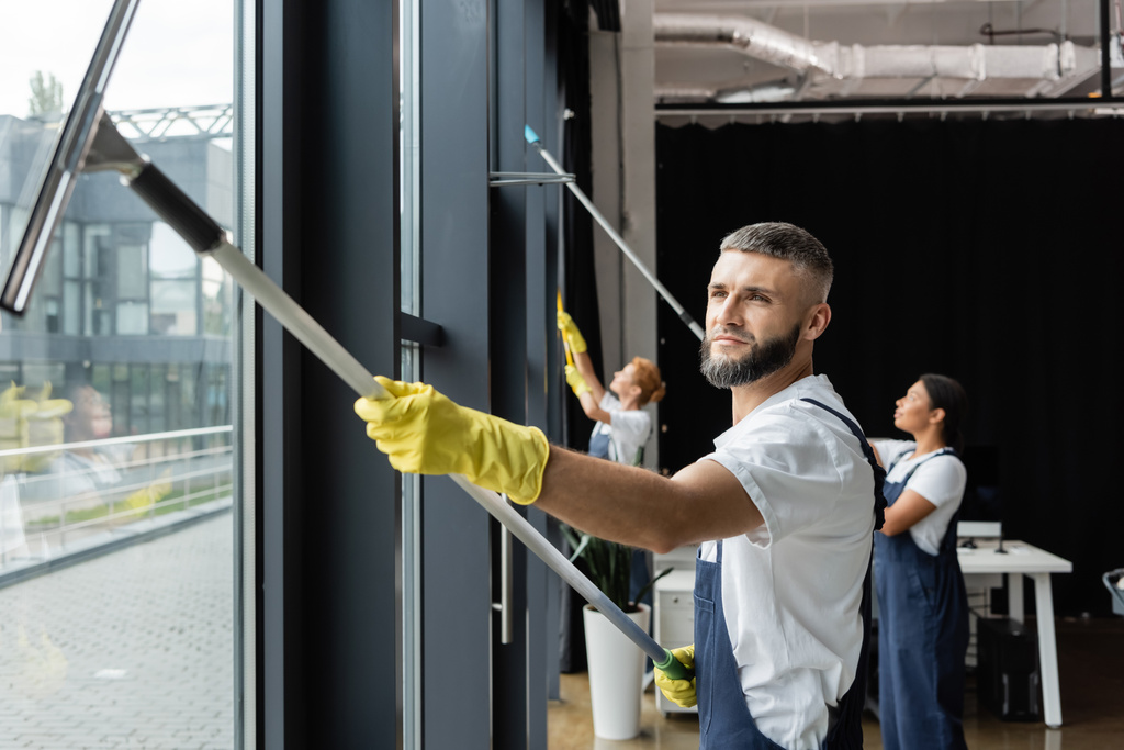 Kauçuk eldivenli sakallı adam ofisteki ırklar arası takımın pencerelerini yıkıyor. - Fotoğraf, Görsel