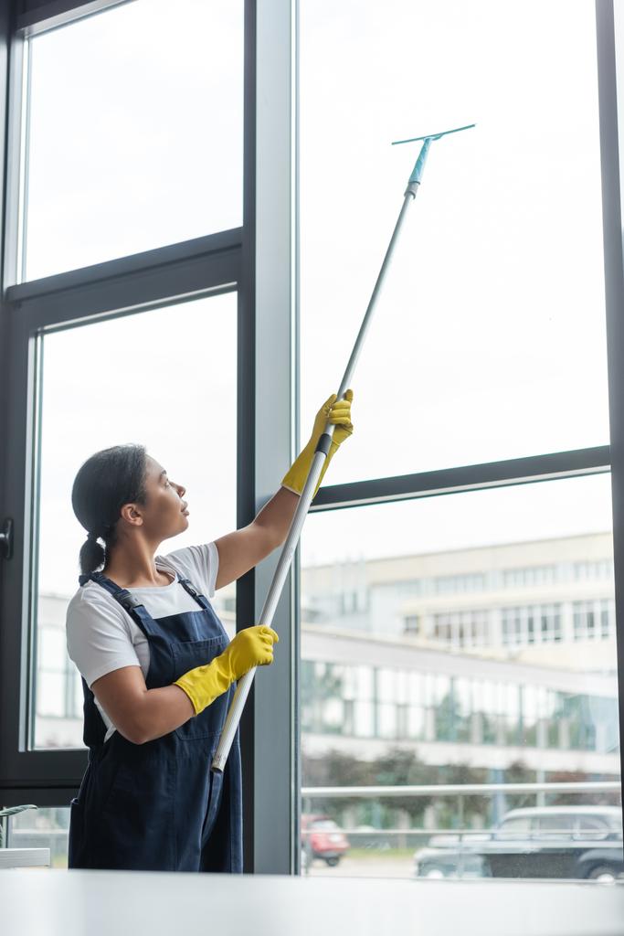 mujer bi-racial en overoles y guantes de goma limpiando grandes ventanas en la oficina - Foto, Imagen