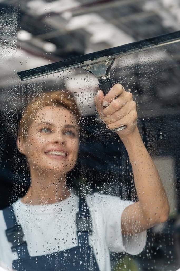 profi takarító ablaktörlővel mosolygós közelében nedves üveg az irodában - Fotó, kép