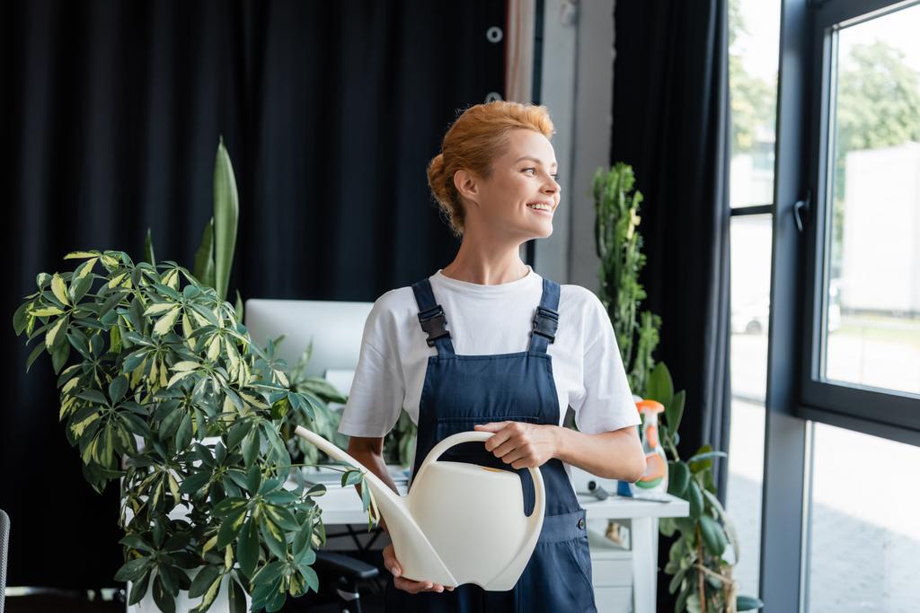 Счастливая женщина с поливной баночкой смотрит в сторону, стоя рядом с растением в офисе - Фото, изображение