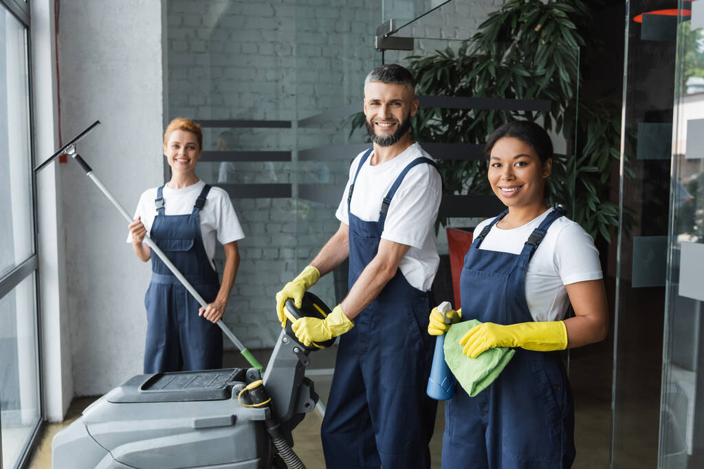 glückliches gemischtrassiges Team professioneller Reinigungskräfte mit Putzutensilien, die im Büro in die Kamera schauen - Foto, Bild