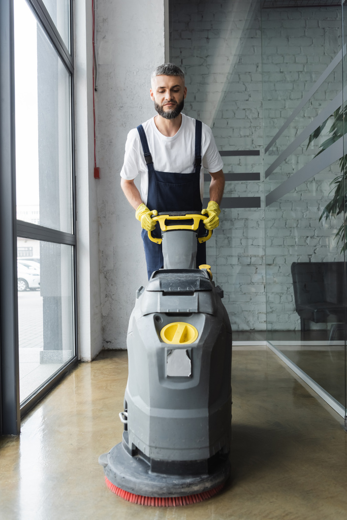 man met baard in overall reinigingsvloer met elektrische scrubbermachine - Foto, afbeelding