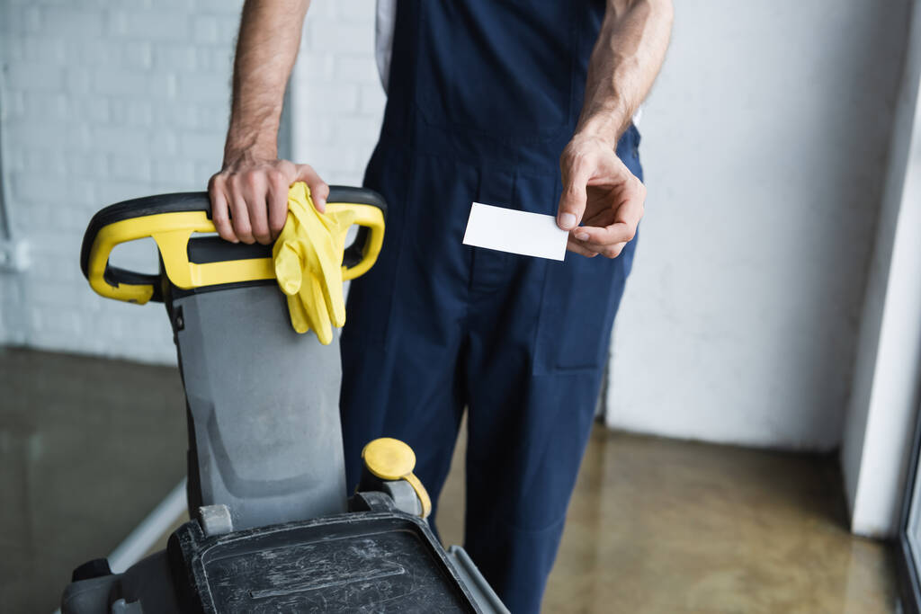 gedeeltelijke weergave van de mens in overalls met blanco visitekaartje in de buurt van vloer schrobzuigmachine - Foto, afbeelding