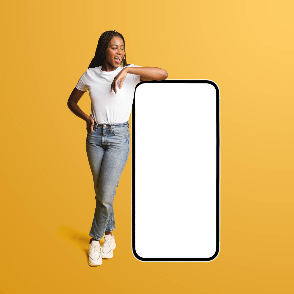 Happy joyful african-american woman leaning on huge smartphone with blank empty screen isolated on yellow, advertising great new mobile app, cheerful teenage girl presenting website, mockup, copyspace - 写真・画像