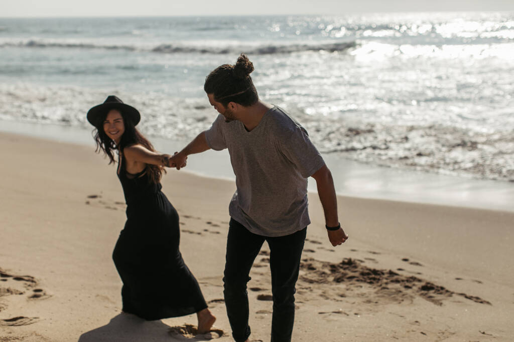 веселая женщина в шляпе и черном платье тянет руку бойфренда на пляже в Португалии - Фото, изображение