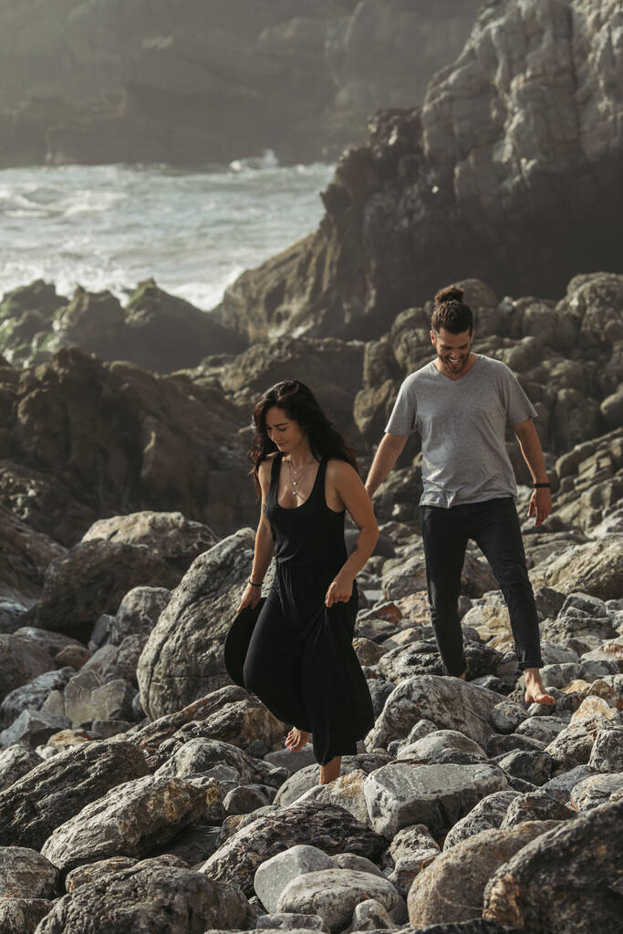 brodaty mężczyzna uśmiecha się podczas spaceru po skałach w pobliżu wytatuowanej dziewczyny w sukience w pobliżu oceanu - Zdjęcie, obraz