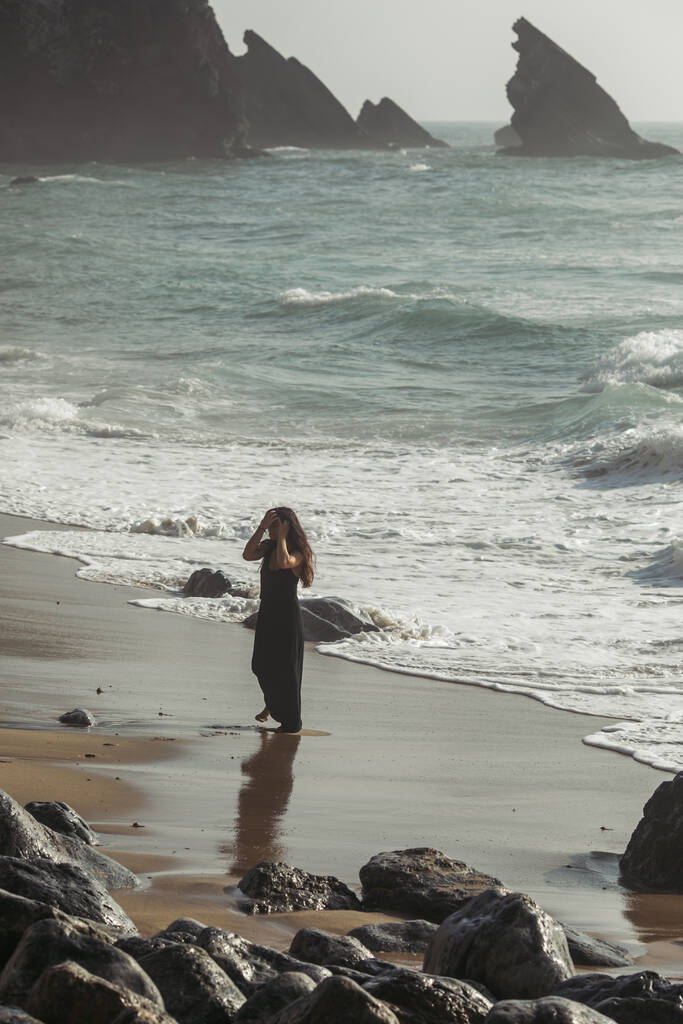 красивая и татуированная женщина в черном платье регулирует волосы, стоя на песчаном пляже в Португалии  - Фото, изображение
