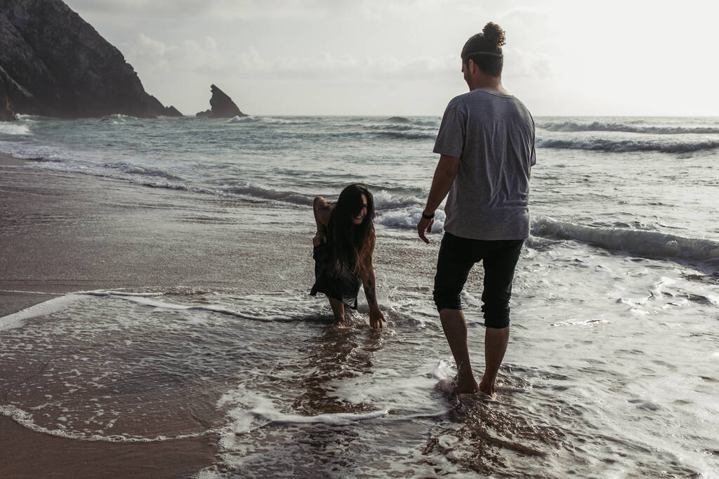 homme barbu regardant petite amie heureuse en robe touchant l'eau de l'océan  - Photo, image