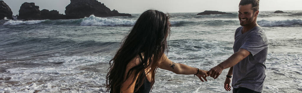бородатый мужчина, держась за руки с татуированной подружкой возле океана, баннер - Фото, изображение