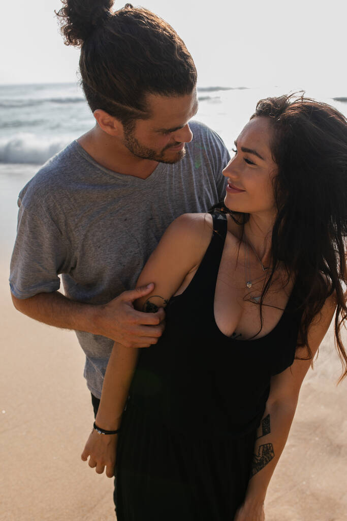 uomo allegro abbracciare fidanzata gioiosa in abito vicino oceano durante le vacanze - Foto, immagini