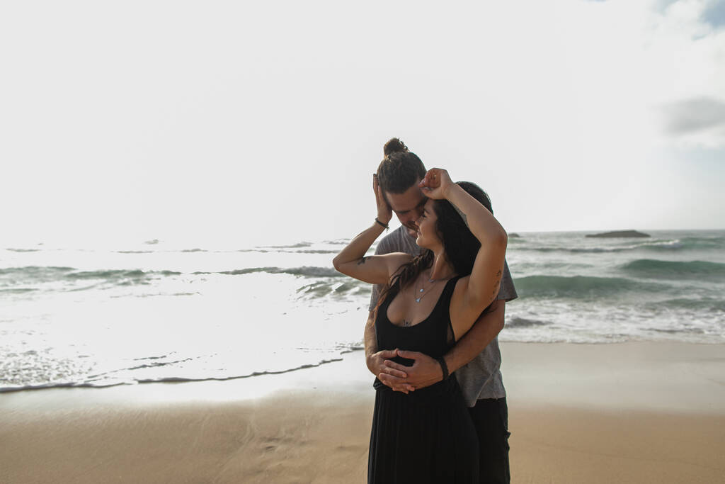 άντρας αγκαλιάζει τατουάζ και χαρούμενη κοπέλα με φόρεμα κοντά στον ωκεανό κατά τη διάρκεια των διακοπών - Φωτογραφία, εικόνα