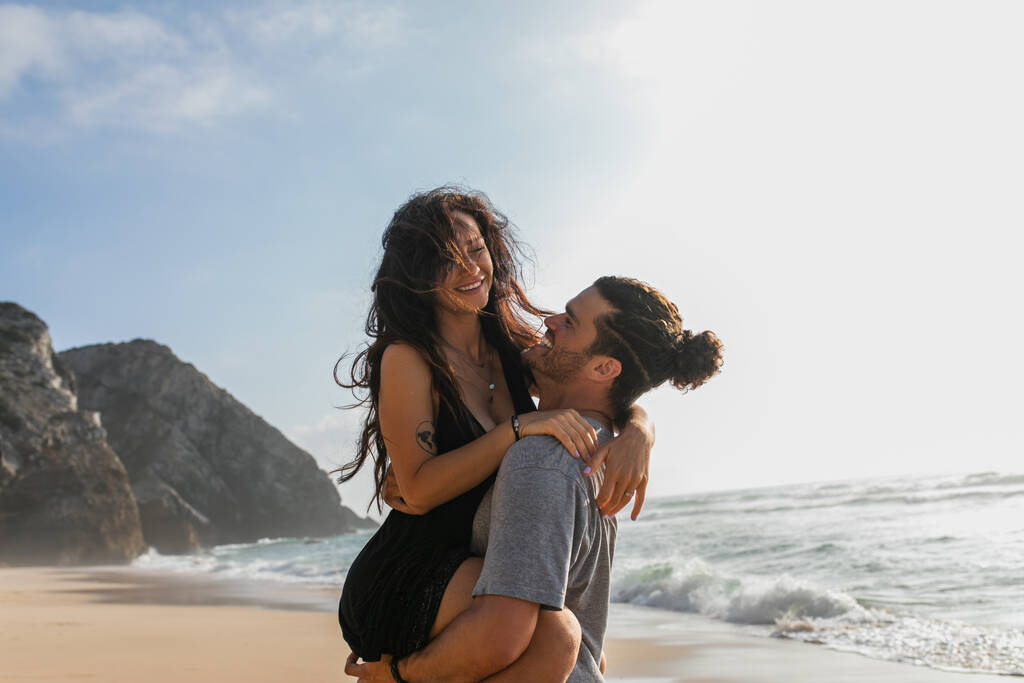 hombre barbudo alegre levantando mujer tatuada y feliz en vestido en la playa cerca del océano  - Foto, imagen