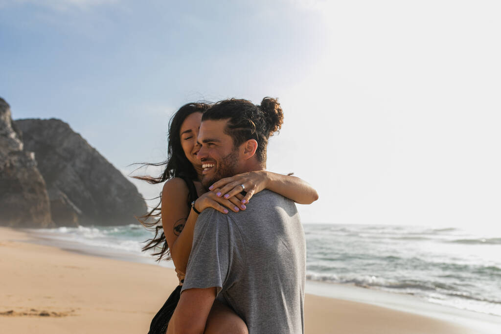 hombre alegre levantando mujer tatuada en la playa cerca del océano  - Foto, Imagen