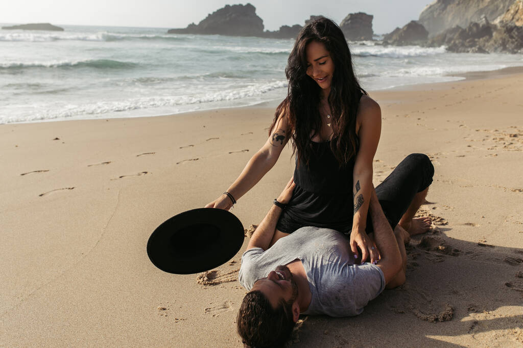 mulher tatuada em chapéu vestido segurando e olhando para o namorado feliz deitado na areia molhada perto do oceano  - Foto, Imagem