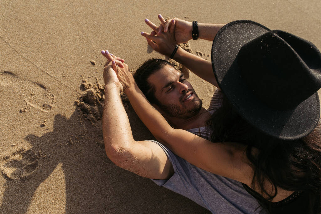 vue de dessus de l'homme barbu tenant la main avec la femme dans le chapeau et couché sur le sable - Photo, image