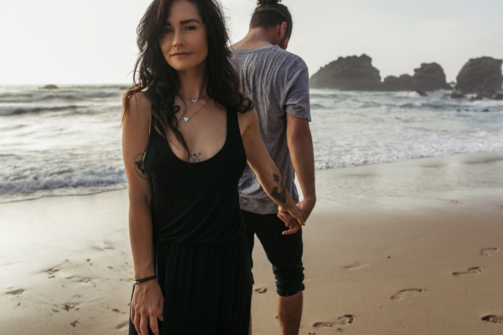 mujer tatuada en vestido y hombre tomados de la mano cerca del océano en la playa en portugal - Foto, Imagen
