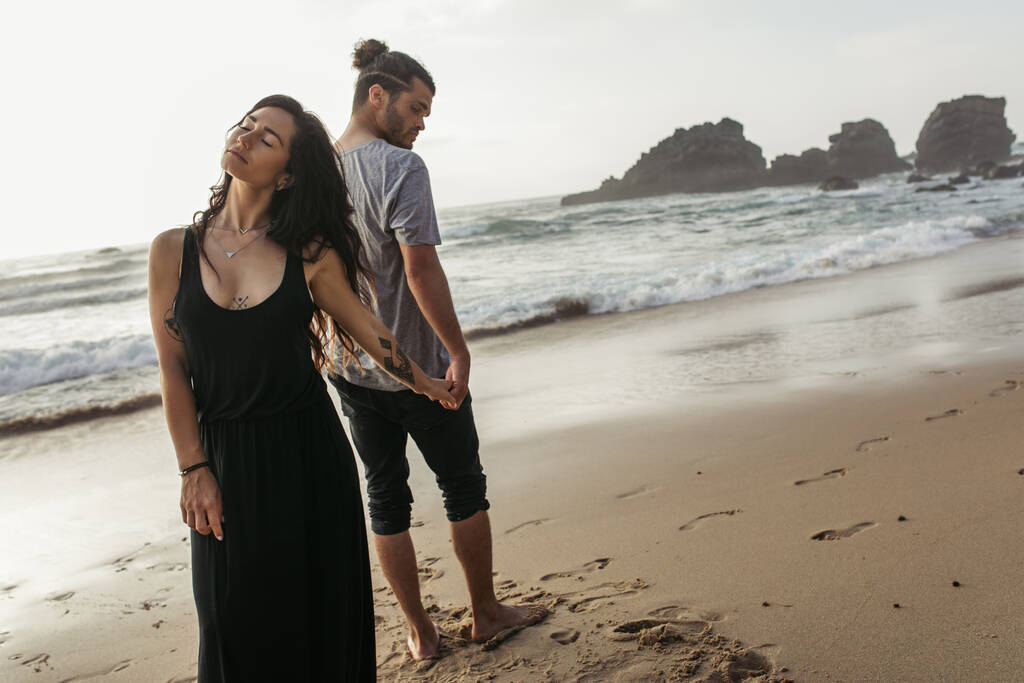 homme barbu et femme tatouée en robe debout sur la plage et tenant la main près de l'océan  - Photo, image