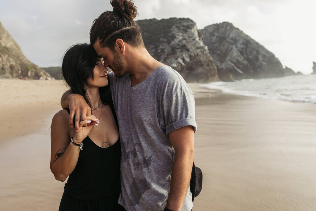 hombre barbudo alegre y mujer tatuada en vestido tomados de la mano cerca del océano  - Foto, imagen