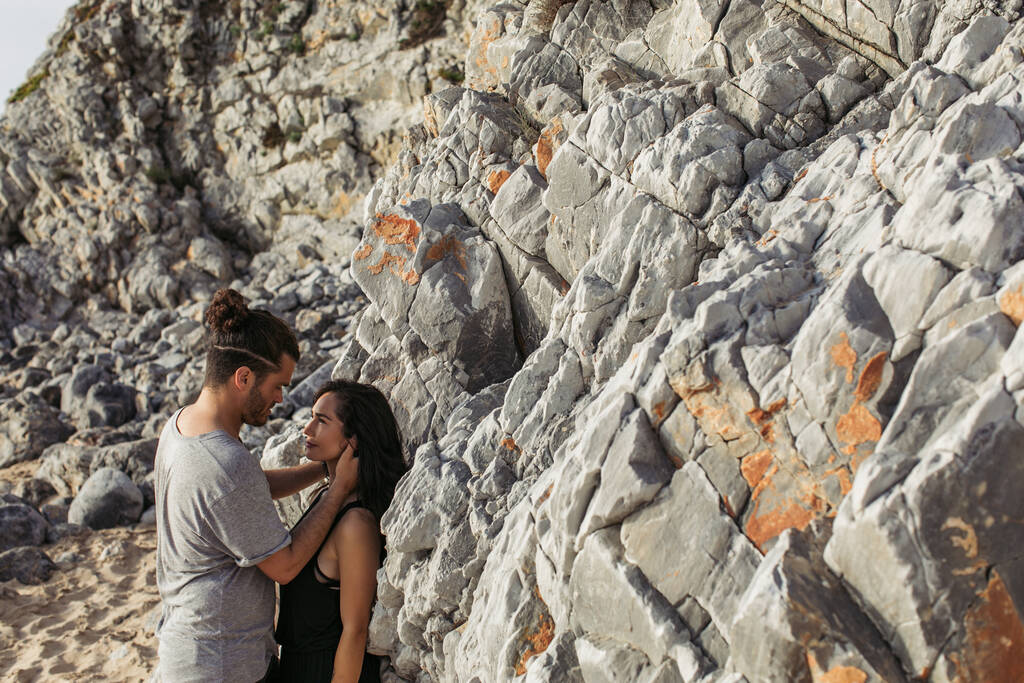side view of bearded man hugging and looking at girlfriend near rocks on beach  - Photo, Image