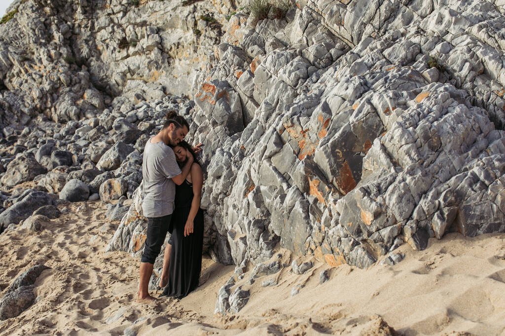 volle Länge des lächelnden Mannes umarmt Freundin im Kleid, während er in der Nähe von Felsen am Strand steht  - Foto, Bild