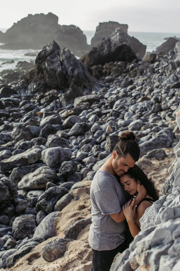 vousatý muž objímání přítelkyně zatímco stojí v blízkosti skály na pláži  - Fotografie, Obrázek