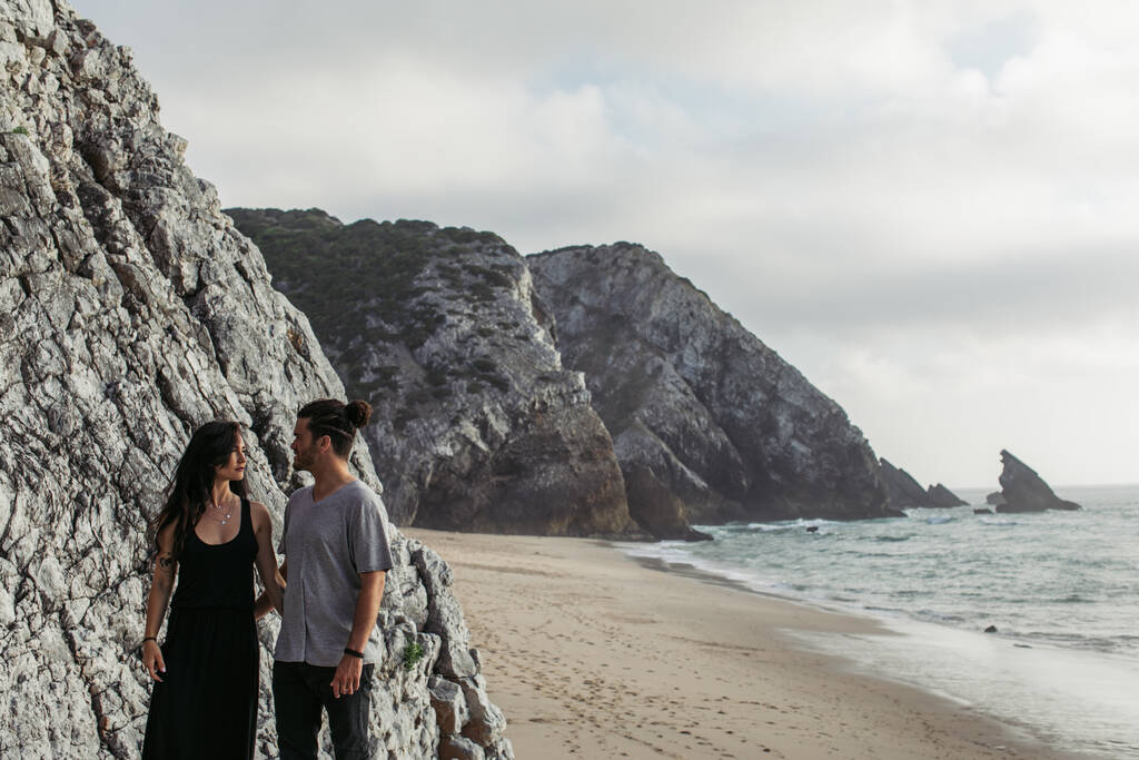 入れ墨女性のドレス抱擁ひげを生やしたボーイフレンドオン岩のビーチ近く海 - 写真・画像
