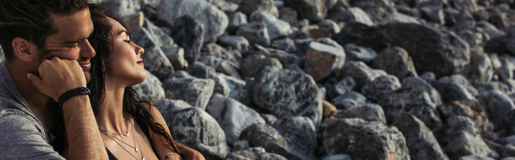 hombre barbudo abrazando mujer tatuada con los ojos cerrados cerca de las rocas en la playa, pancarta - Foto, imagen
